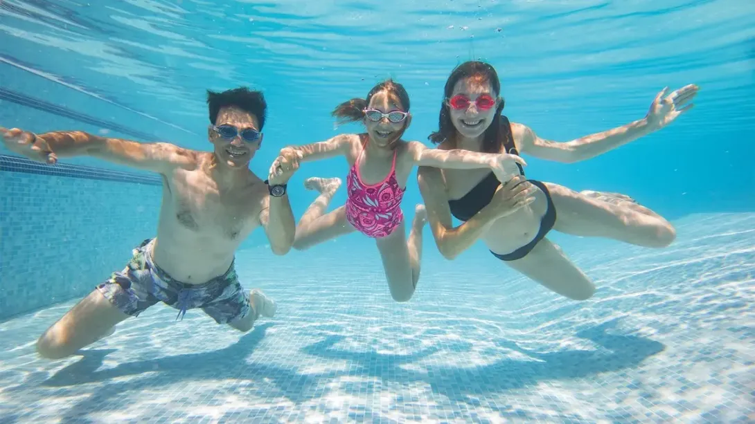 Tres personas bajo el agua en piscina, tomadas de la mano y usando gafas de natación.